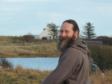 A white man with long hair tied back, looks back towards the camera from over his shoulder. He is in a rural location with a loch and grasses in the background. 