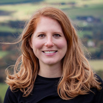 A red-haired white woman stands smiling in the photo, with a view of a valley behind her